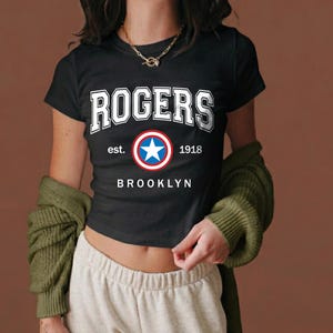 Peut inclure: T-shirt noir avec le mot "ROGERS" en lettres blanches. En dessous, un embl&egrave;me circulaire avec une &eacute;toile bleue, entour&eacute;e de rouge et de blanc. Le texte "est. 1918 BROOKLYN" est imprim&eacute; en dessous.