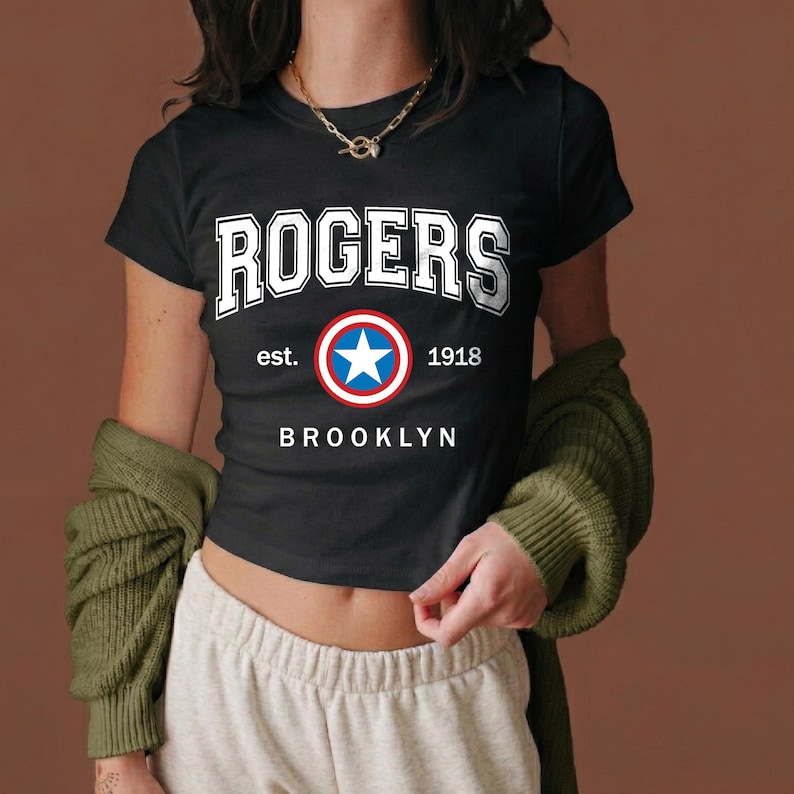 Peut inclure: T-shirt noir avec le mot "ROGERS" en lettres blanches. En dessous, un embl&egrave;me circulaire avec une &eacute;toile bleue, entour&eacute;e de rouge et de blanc. Le texte "est. 1918 BROOKLYN" est imprim&eacute; en dessous.