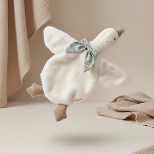 May include: A plush white goose-shaped baby comforter with a light green floral bow. The comforter has beige feet and beak. Soft beige blankets are in the background.