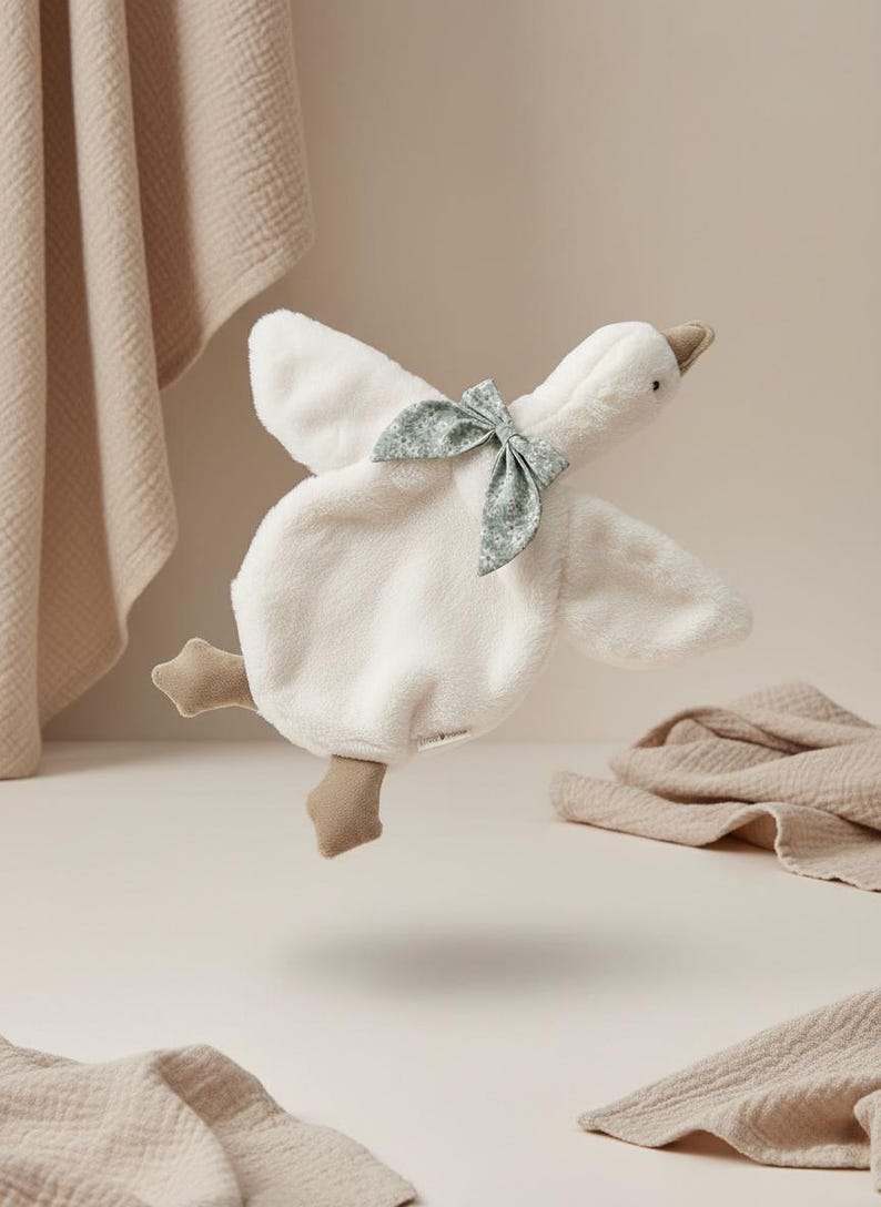 May include: A plush white goose-shaped baby comforter with a light green floral bow. The comforter has beige feet and beak. Soft beige blankets are in the background.