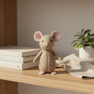 May include: A tan-colored stuffed mouse with pink ears and feet stands on a wooden shelf. The plush toy has a stitched face with whiskers. A stack of books and a small potted plant are in the background.