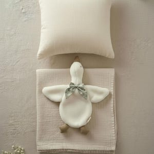 May include: A cream-colored pillow and a soft, light pink blanket with a white goose-shaped plush toy. The toy has a green floral bow and brown feet. The items are arranged on a neutral-toned surface.