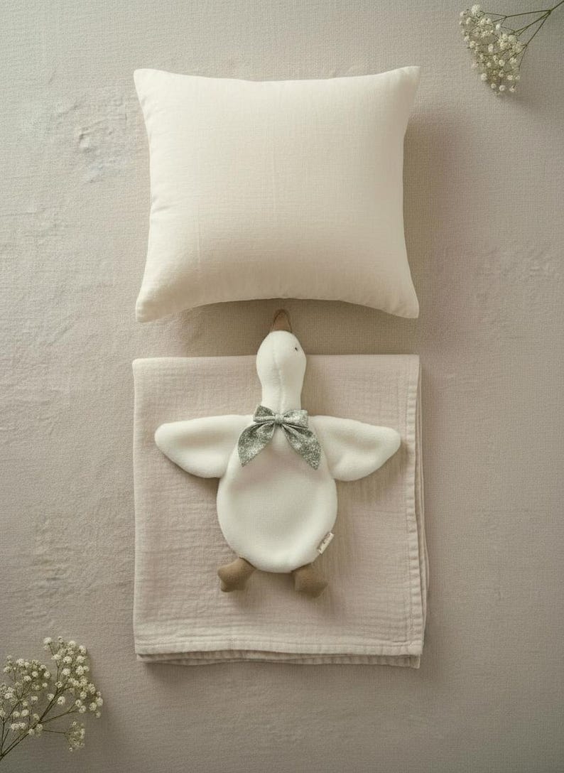 May include: A cream-colored pillow and a soft, light pink blanket with a white goose-shaped plush toy. The toy has a green floral bow and brown feet. The items are arranged on a neutral-toned surface.
