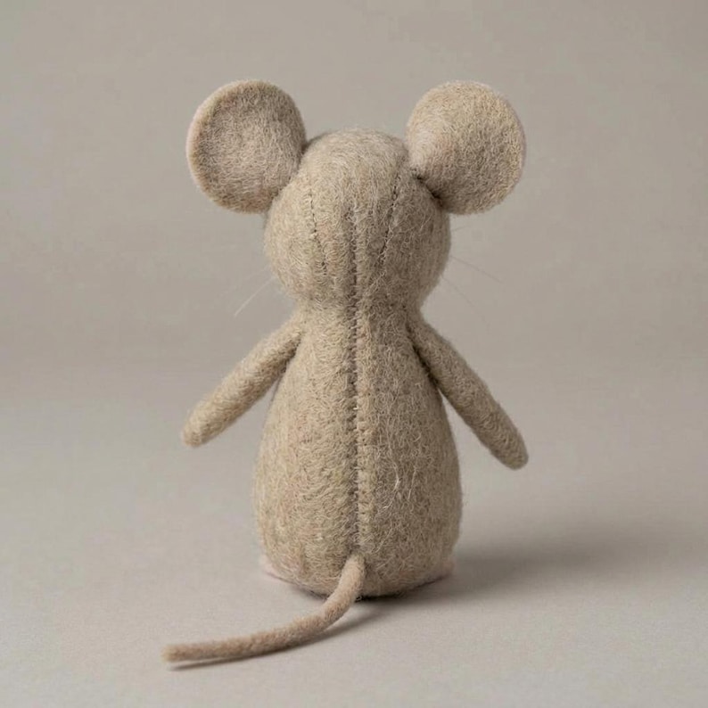 May include: A handmade, beige felt mouse doll viewed from the back. The doll has large, rounded ears, a long tail, and small arms. The stitching is visible along the seams. The mouse is a soft, neutral color, and the background is a solid, light beige.
