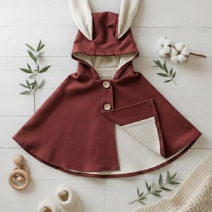 Puede incluir: Una capa con capucha burdeos con orejas de conejo, dos botones de madera y un interior color crema. La capa se combina con patucos de bebé color crema, un juguete de madera y una manta de punto sobre una superficie de madera blanca.