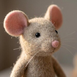 May include: A handcrafted felt mouse figurine. The mouse has a light brown body, large pink ears, a pink nose, and black bead eyes. The whiskers are thin and white. The mouse is a charming, handmade decorative item.