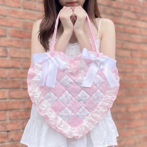May include: A pink heart-shaped tote bag with white bows and lace trim. The bag features a quilted design with pink and white checkered squares and floral print squares. The tote has pink handles.