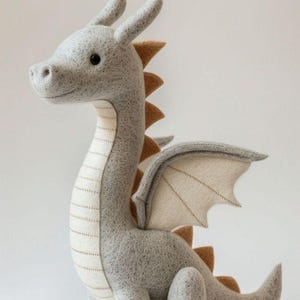 May include: A soft grey dragon toy with tan details. The dragon features a long neck, small wings, and a tail. The dragon's belly is cream-coloured with a stitched design. The dragon is sitting and has a friendly expression.