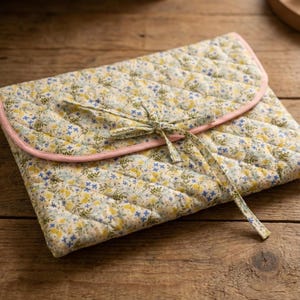 Puede incluir: Clutch acolchado con estampado floral. La tela presenta un fondo amarillo claro con flores azules y amarillas, un ribete rosa y un patrón de diamantes. Se ata con una cinta a juego.