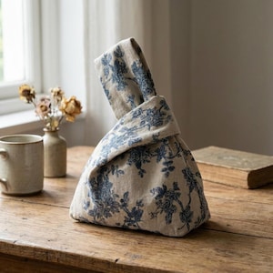 Puede incluir: Una bolsa de tela beige con un estampado floral azul. La bolsa está anudada en la parte superior, creando un asa. La bolsa está sobre una mesa de madera. Una taza, un jarrón y un libro están en el fondo.