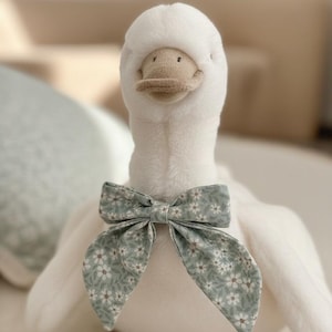 May include: A plush white goose toy with a light brown beak and feet. The goose wears a bow tie with a floral pattern in shades of green and white. The toy is soft and fluffy, perfect for a child's room.