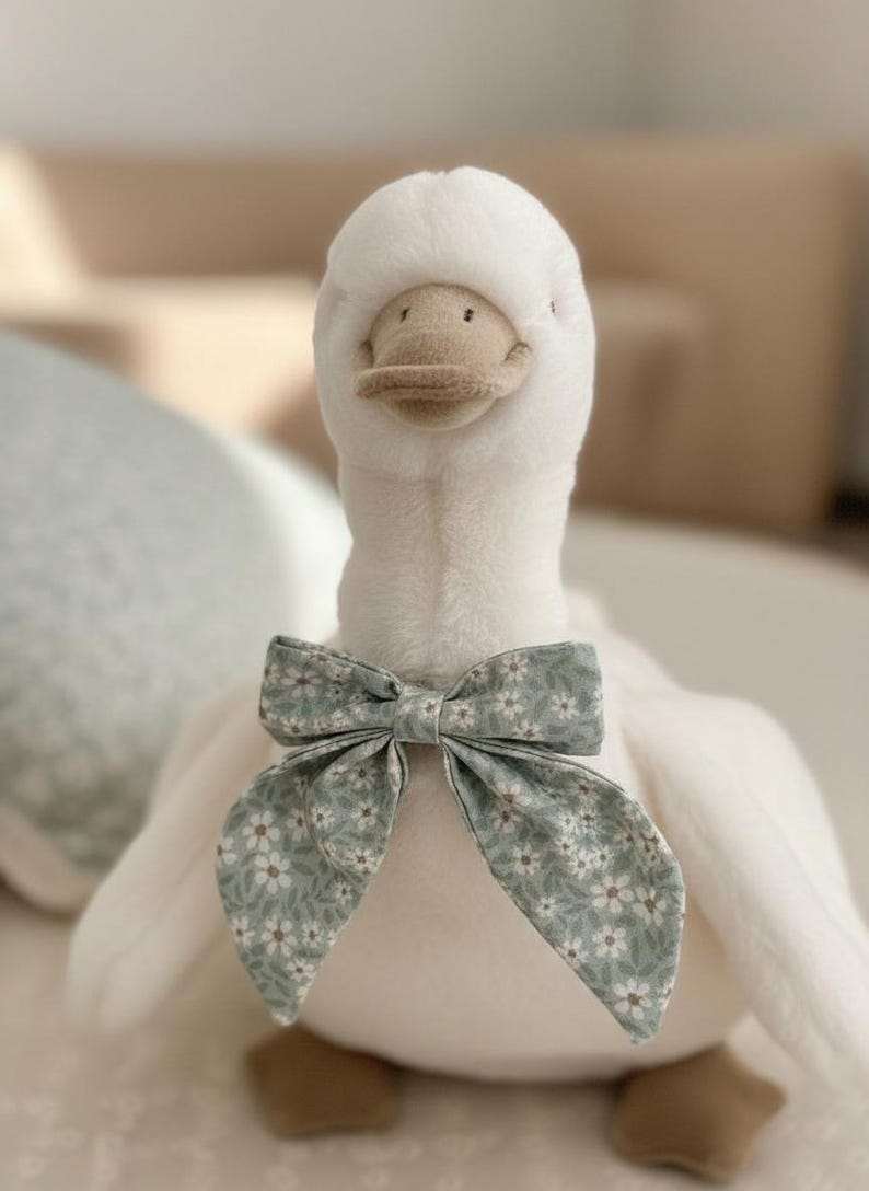May include: A plush white goose toy with a light brown beak and feet. The goose wears a bow tie with a floral pattern in shades of green and white. The toy is soft and fluffy, perfect for a child's room.