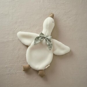 May include: A plush white goose toy with outstretched wings and a gray patterned bow tie. The goose has tan feet and a tan beak. The toy is lying on a neutral-colored surface.