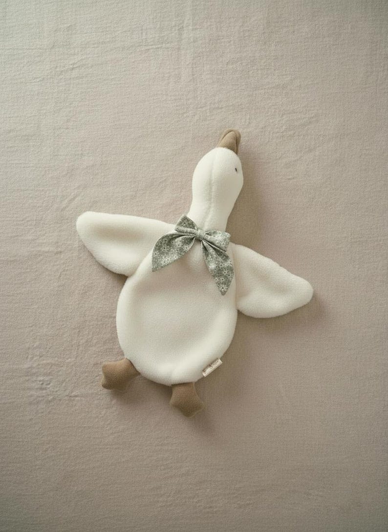 May include: A plush white goose toy with outstretched wings and a gray patterned bow tie. The goose has tan feet and a tan beak. The toy is lying on a neutral-colored surface.