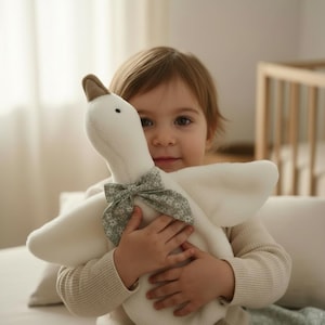 May include: A soft, white plush goose toy with a brown beak and feet, and a floral bow. The toy is being held by a child wearing a cream-colored sweater. The background is a soft, neutral color.