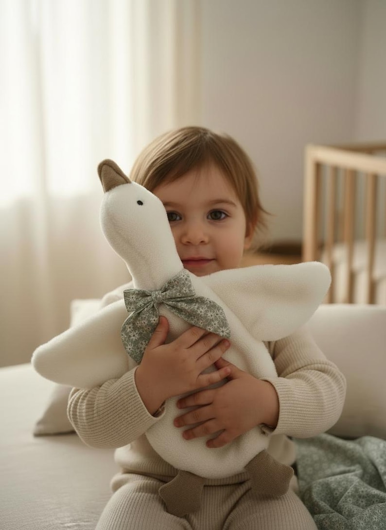 May include: A soft, white plush goose toy with a brown beak and feet, and a floral bow. The toy is being held by a child wearing a cream-colored sweater. The background is a soft, neutral color.