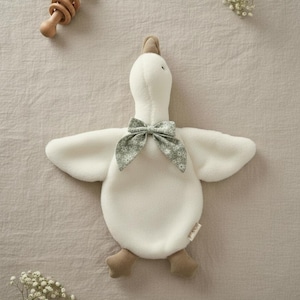 May include: A plush white goose toy with a green floral bow tie, brown feet, and a brown head. A wooden rattle is in the upper left corner. The toy is on a beige surface, with sprigs of white flowers.