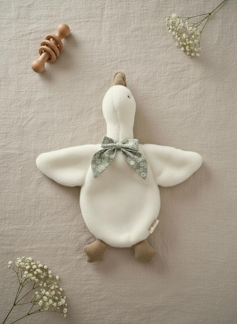 May include: A plush white goose toy with a green floral bow tie, brown feet, and a brown head. A wooden rattle is in the upper left corner. The toy is on a beige surface, with sprigs of white flowers.