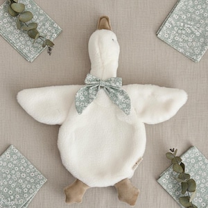 May include: A plush white goose-shaped baby comforter with a floral bow tie and brown feet. The comforter is surrounded by four floral print napkins and sprigs of greenery, all on a neutral background.