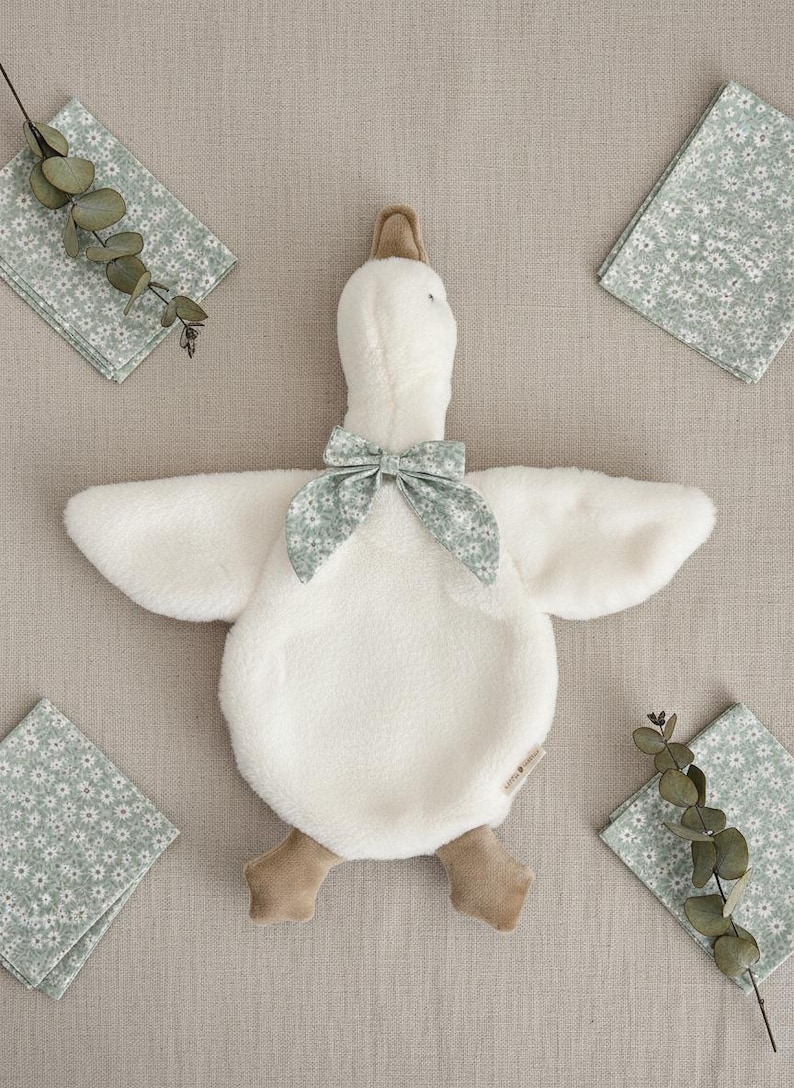 May include: A plush white goose-shaped baby comforter with a floral bow tie and brown feet. The comforter is surrounded by four floral print napkins and sprigs of greenery, all on a neutral background.