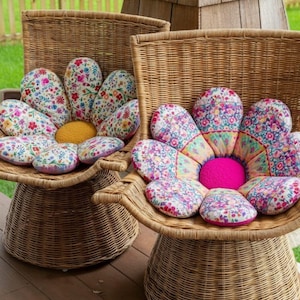 Może przedstawiać: Dwa wiklinowe krzesła z poduszkami w kształcie kwiatów. Poduszki mają kwiatowy wzór w odcieniach różu, błękitu i żółci. Środek jednej poduszki jest jasno różowy, a drugiej żółty. Krzesła stoją na drewnianym tarasie.