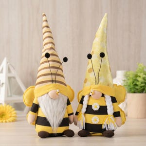 May include: Two plush bee gnomes. One gnome has a striped brown and tan hat, the other has a yellow dotted hat. Both have yellow and black striped bodies, white beards, and bee antennae. They are decorated with small daisies.