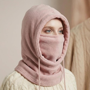 Peut inclure: Une cagoule en polaire rose clair avec capuche et couvre-visage. La cagoule a un cordon de serrage et couvre le cou, le visage et la tête. La personne sur l'image porte un pull crème.