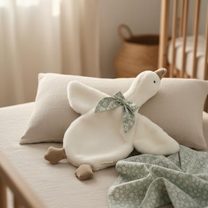May include: A soft white goose toy with a green floral bow and brown feet rests on a bed with a matching blanket. A beige pillow and a wooden cot are in the background.