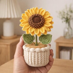May include: A handcrafted crochet sunflower in a cream-colored pot. The sunflower has yellow petals and a brown center. Green leaves and a stem support the flower. The pot has a scalloped edge.