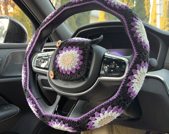 May include: A crocheted steering wheel cover and matching accessory in black, white, and purple. The cover features a floral pattern and is installed on a car's steering wheel. The accessory is attached with wooden buttons.