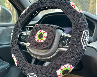 May include: Black crochet steering wheel cover with white spiderwebs and red, green, and white eye designs. A matching cover is on the center of the steering wheel. The car interior is visible in the background.