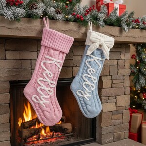 May include: Two cable-knit Christmas stockings, one pink and one blue, hang above a fireplace. Each stocking has the name "Kelly" embroidered in white script. The pink stocking has a matching pink cuff, while the blue stocking has a white cuff with a bow.