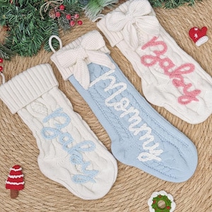 Puede incluir: Tres calcetines navideños de punto trenzado en blanco y azul claro, con lazos en la parte superior. Un calcetín dice "Mommy" en escritura blanca, y dos dicen "Baby" en escritura rosa y azul claro. Decoración festiva navideña.