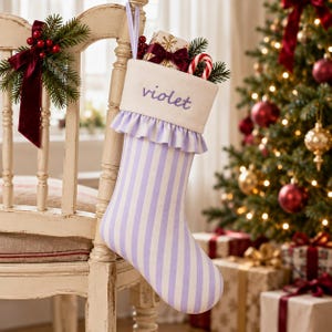 Op de afbeelding: Een kerstkous met een lichtpaars en wit verticaal gestreept patroon, voorzien van een ruche en een witte boord waarop de naam violet is geborduurd. De kous is gevuld met een zuurstok en een klein cadeaudoosje met een donkerrode strik.