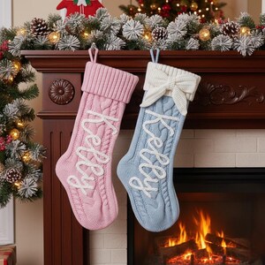 May include: Two cable-knit Christmas stockings, one pink and one blue, hang from a fireplace mantel. Each stocking has the name "Kelly" embroidered in white script. The blue stocking has a white bow at the top.