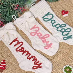 May include: Three white cable-knit Christmas stockings with bows and the words "Mommy," "Baby," and "Daddy" in red, pink, and green, respectively. Each stocking has a loop for hanging. Festive holiday decor.