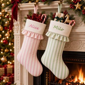 May include: Two Christmas stockings with white cuffs, one featuring pink stripes and the name Alice in pink embroidery, the other with green stripes and the name Milo in green embroidery. Both have ruffled trim and are filled with holiday decor.