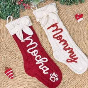 May include: Two cable-knit Christmas stockings, one red and one white, with white bows and the names "Mocha" and "Mommy" embroidered in white and red, respectively. A paw print is on the red stocking. Festive holiday decor.
