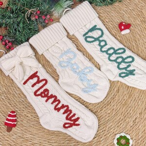 May include: Three cream-colored Christmas stockings with cable knit design. One stocking has a red bow, another has the word "Mommy" in red, "Baby" in light blue, and "Daddy" in green. Small decorative items are scattered around the stockings.
