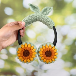 May include: Crocheted earmuffs with sunflower designs. The ear covers are yellow, orange, and brown, with a green border. The headband is green with two small, leaf-shaped accents. The ear muffs are held by a person.