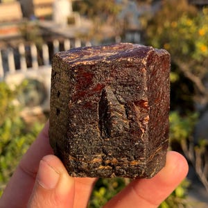 May include: A dark red, cube-shaped mineral specimen with a rough, textured surface. The stone has a deep, rich colour and appears to be a natural, uncut gemstone. The background is blurred, suggesting an outdoor setting.