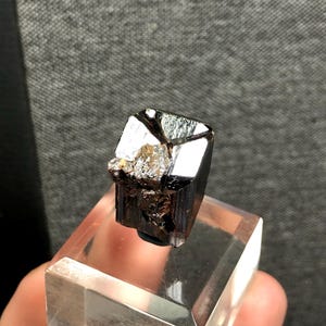 Könnte beinhalten: Dunkles, glänzendes schwarzes Mineralexemplar mit kristalliner Struktur, montiert auf einem transparenten Acryl-Präsentationsständer. Das Mineral hat reflektierende Oberflächen und eine raue, strukturierte Oberfläche. Der Hintergrund ist ein neutrales Grau.