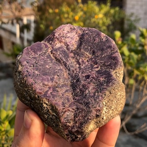 Puede incluir: Una gema en bruto, sin cortar, de color púrpura intenso, que muestra texturas y variaciones naturales. La piedra tiene una forma redondeada y una mezcla de tonos oscuros y claros, lo que sugiere una estética cruda y terrosa.