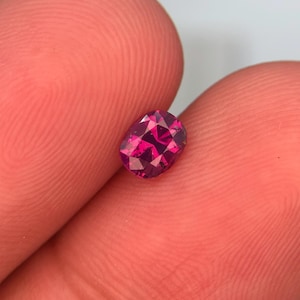 Può includere: Una gemma sfaccettata, di forma ovale, color magenta intenso. La gemma è tenuta tra due dita, mettendo in risalto la sua taglio brillante e il colore vibrante. Le sfaccettature della gemma riflettono la luce. Adatta per la creazione di gioielli.