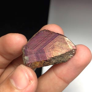 May include: A hand holds a raw mineral specimen. The stone is a deep purple colour with iridescent, metallic streaks and a rough, textured surface. The edges are a mix of brown and gold.