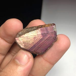 Pode incluir: Uma mão segurando um espécime mineral bruto e natural. A pedra tem um exterior áspero e texturizado com um brilho iridescente roxo profundo e dourado. O interior do mineral exibe uma estrutura cristalina em camadas.