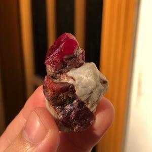 以下が含まれることがあります： 深い赤色の原石ルビー。白と灰色のマトリックスに部分的に埋め込まれています。ルビーの表面は光を反射し、その結晶構造を示しています。天然の鉱物標本です。