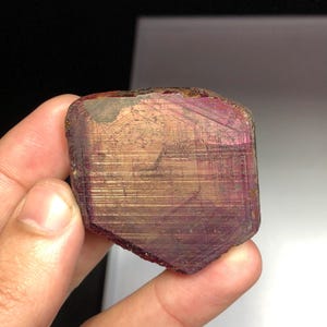 May include: A faceted, rectangular gemstone with a mix of colors, including deep red, pink, and gold. The stone has a rough, textured surface and appears to be held by a person's hand. The gem has a natural, raw appearance.