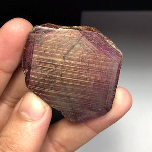 Può includere: Una gemma naturale grezza, di colore bordeaux intenso e struttura cristallina. La pietra ha una superficie ruvida e irregolare con striature visibili e una sottile lucentezza dorata. La gemma è tenuta in una mano.