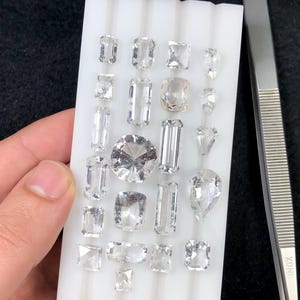 Può includere: Una collezione di gemme tagliate di varie forme, tra cui smeraldo, a goccia e rotonde, esposte su un vassoio bianco. Le gemme sono chiare e scintillanti, con sfaccettature che catturano la luce. Visibile una pinzetta in acciaio inossidabile.